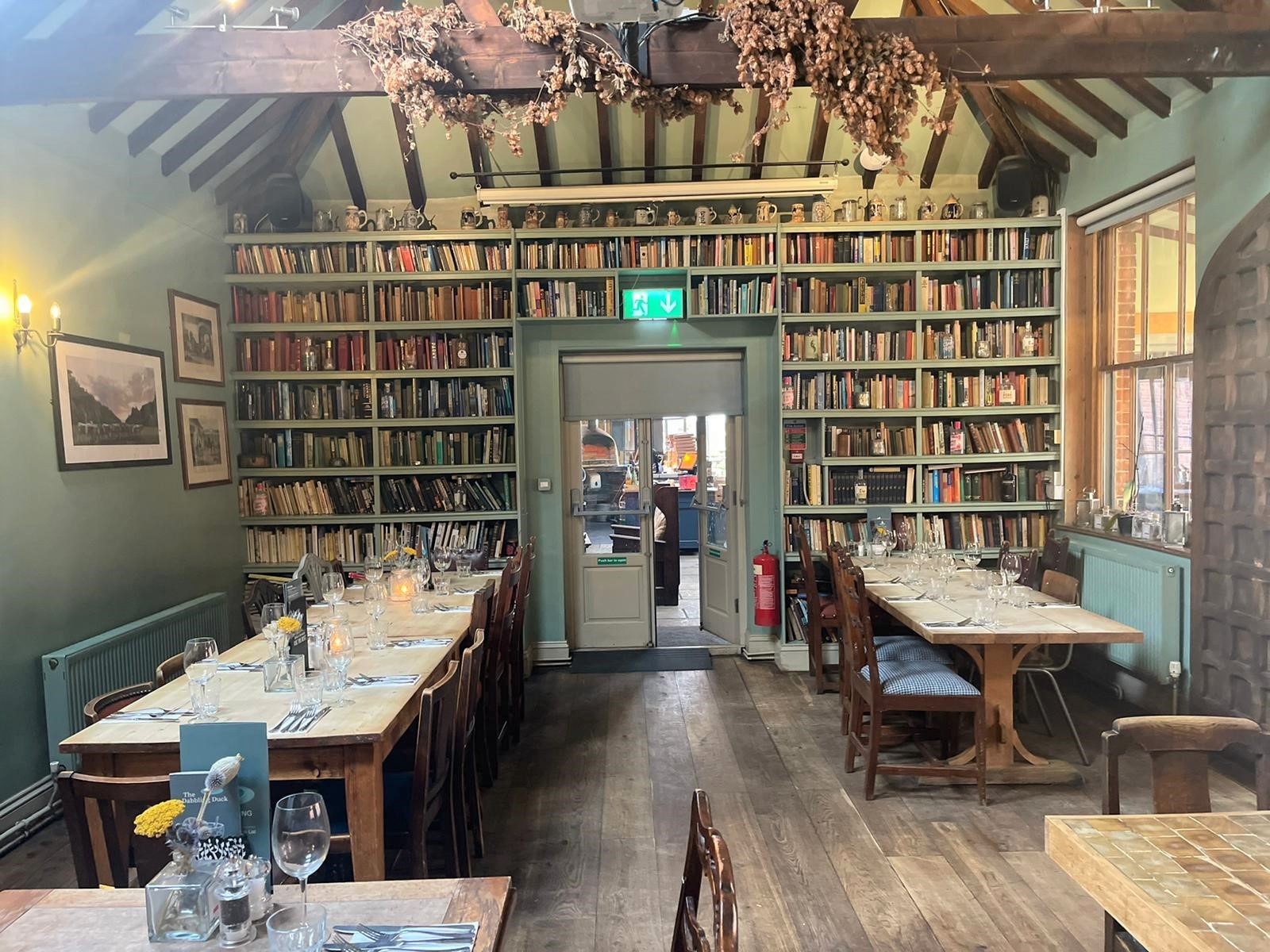 The interior of a restaurant with rustic decor, wooden dining table and chairs, there are wooden beams decorated in flowers and the back wall is lined with vintage books