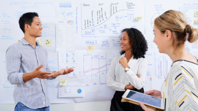 3 people having a conversation in front of a wall that has large pieces of paper attached to it. The papers have charts, figures, and the steps of a marketing plan on them