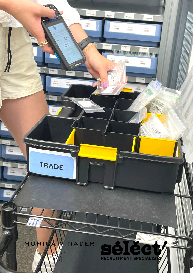 A box is filled with small bags containing pieces of jewellery, there us a person holding to of the bags over the box scannign barcodes with a handheld device