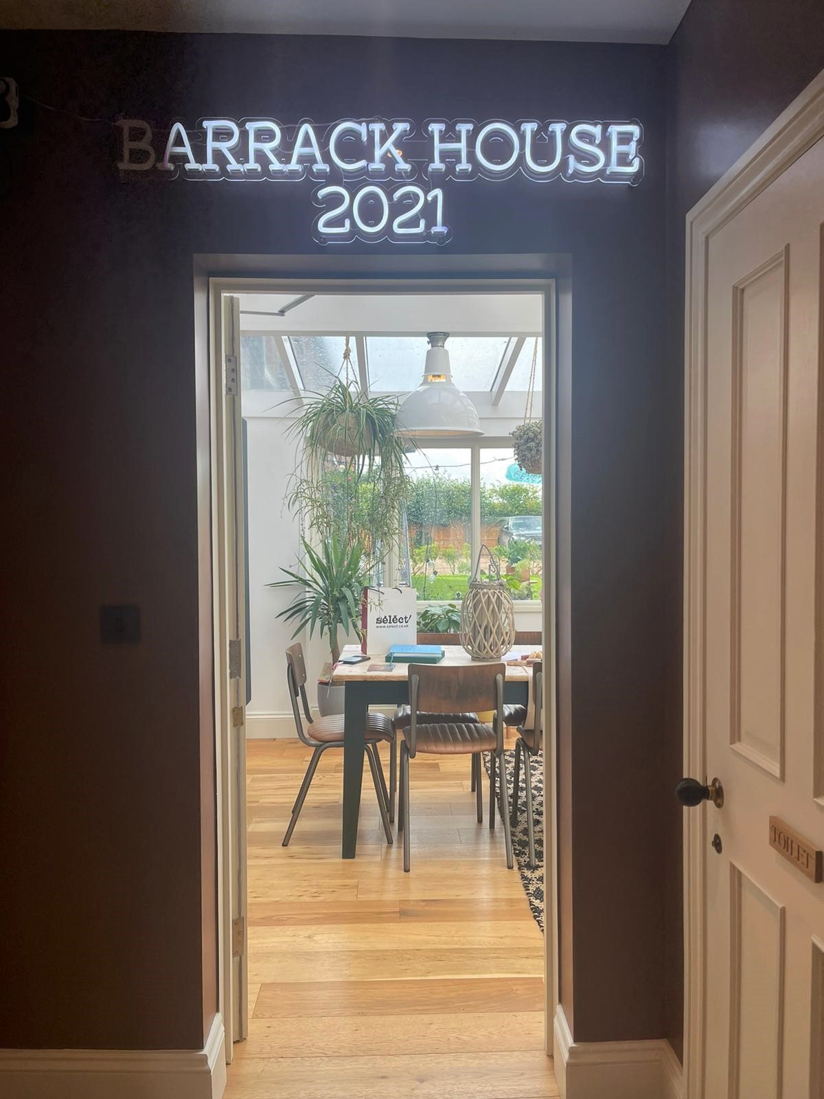 An open doorway with a view of a restaurant dining room, hanging above the dining table and chairs are plants and a rustic pendant light, above the doorway the sigh reads Barrack House 2021