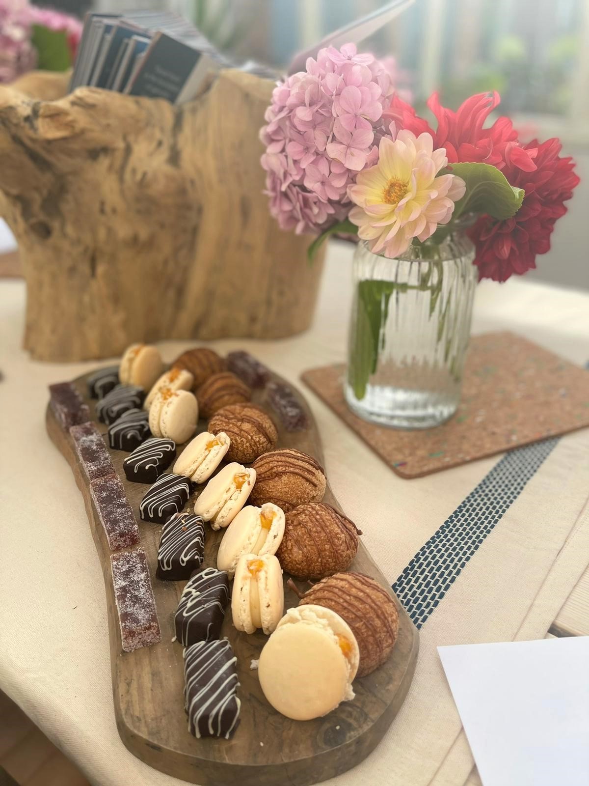 A wooden plate with rows of cakes on it which sits in front of a glass case filled with pink and purple flowers