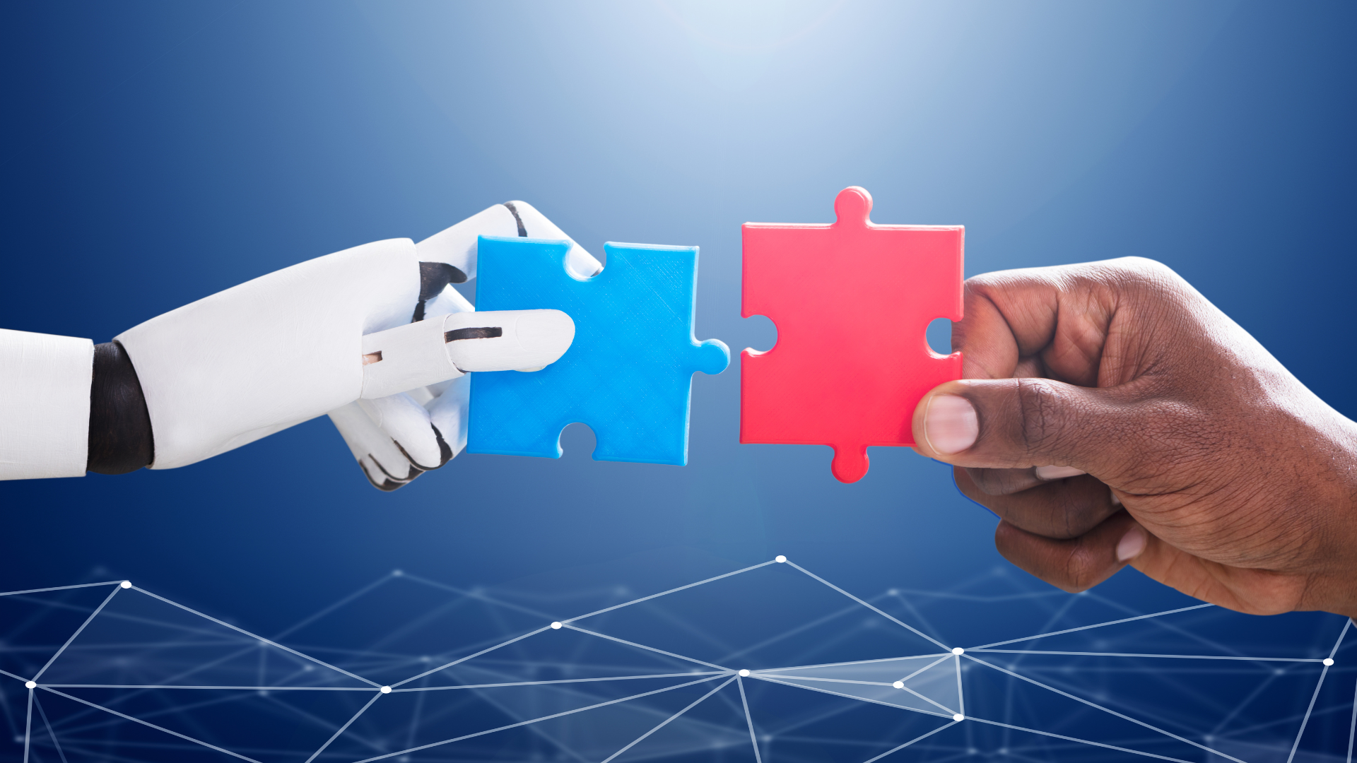 A robot hand holding a blue jigsaw piece, connecting it to a red jigsaw piece that is held by a human hand