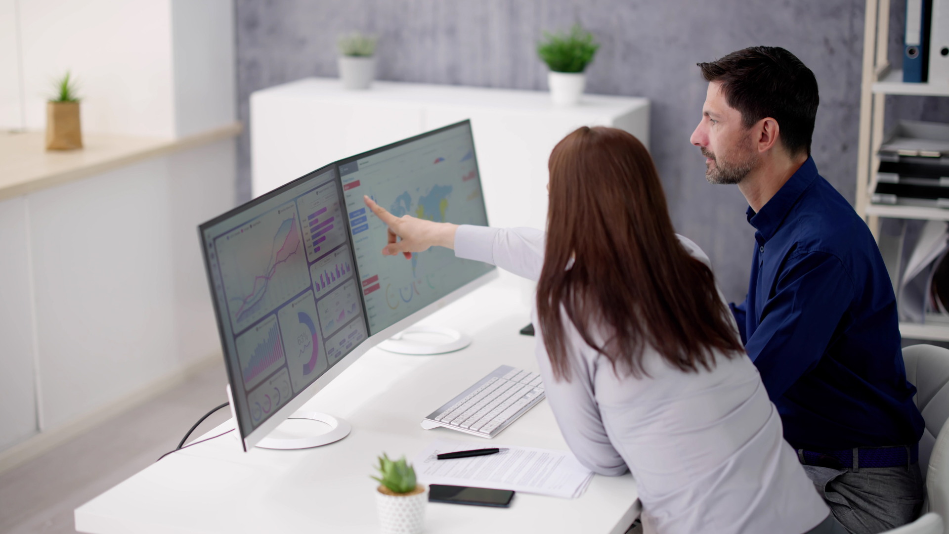 A man and a woman looking at marketing analytics on two computer screens on a white desk. The analytics show growth trends