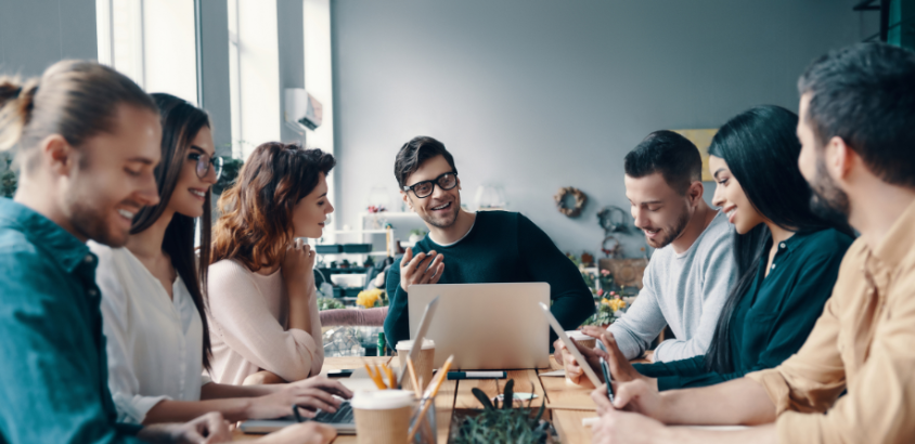 An in-house marketing team having a meeting at a wooden table. People are using different digital devices and having a discussion