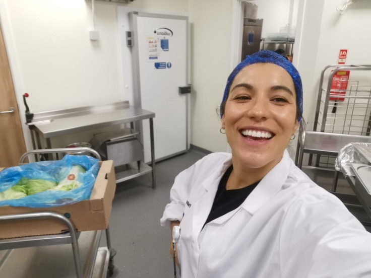 A woman wearing a white chefs uniform and a blue hair, she is standing in a professional kitchen smiling at the camera