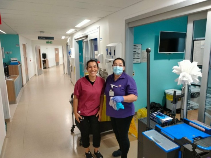 Two women standing in a hospital holding cleaning equipment, the woman on the right is wearing disposable gloves and facemask