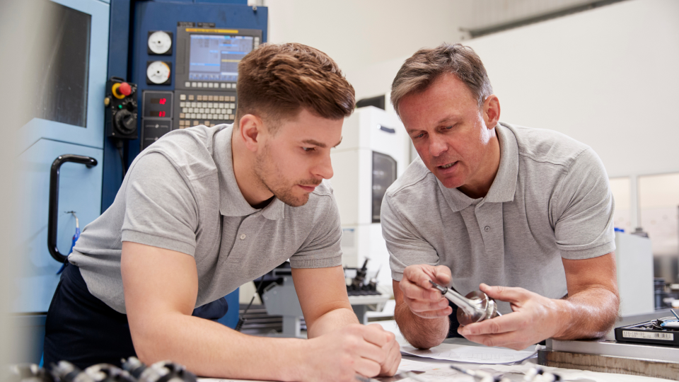 An image of two engineering workers in a workshop discussing components. They are both wearing grey polo-shirts, and there are CNC machines behind them.