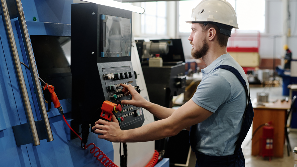 An image of a worker using a CNC machine in a factory. He is pressing buttons on a control panel, and is wearing a hard-hat.