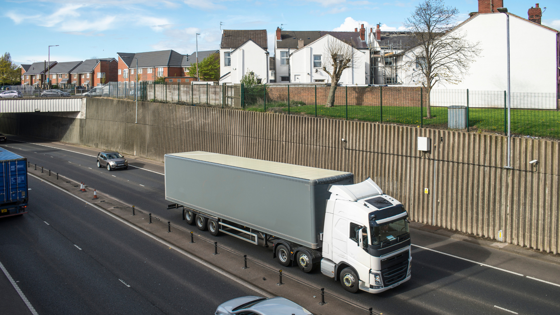 An image of a delivery lorry driving on a dual-carriageway road. There are modern houses above the road.