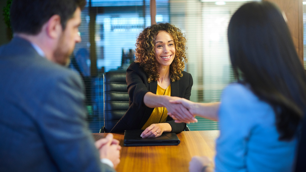 An image of a woman shaking a person's hand after a job interview. She is sat across a wooden table from two people dressed in suits.
