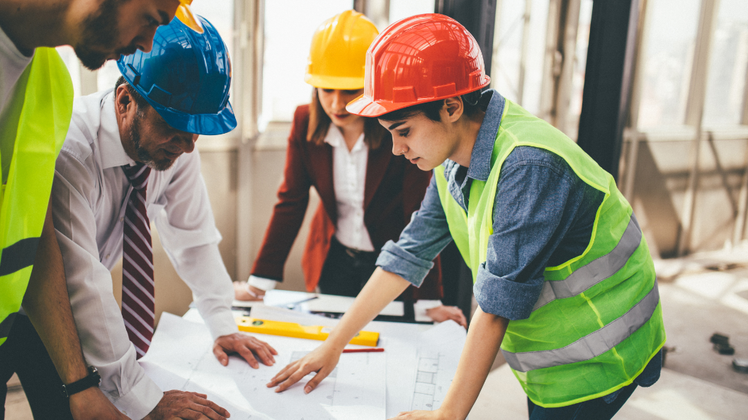 An image of people looking at construction plans on a building site. Two people are wearing high visibility vests, two are wearing smart office wear.