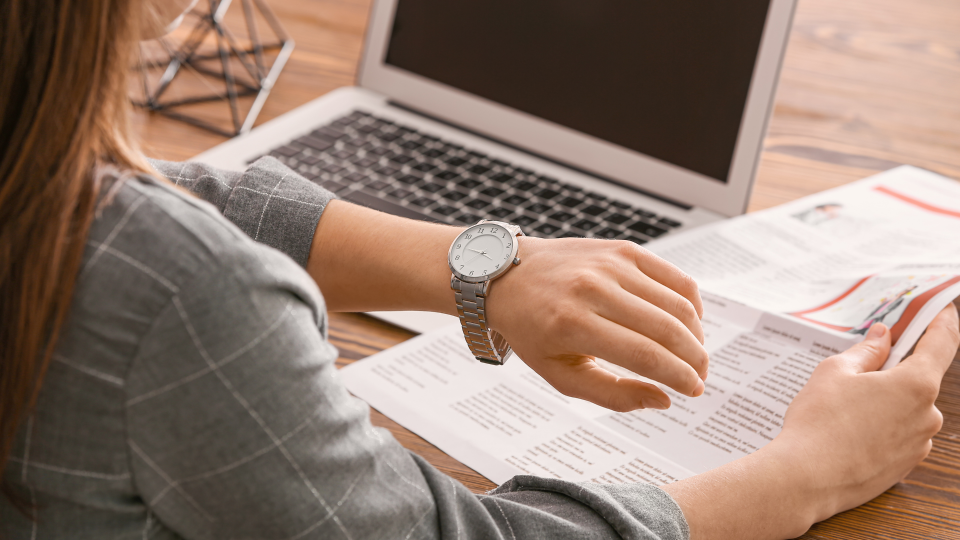 An image of a woman at work checking her watch. She is sat at a wooden desk that has a laptop and papers on it.