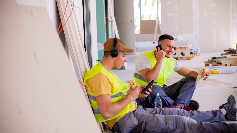 An image of two construction workers taking a break. They are both eating sandwiches, one is listening to music, while the other is making a call on a mobile phone.