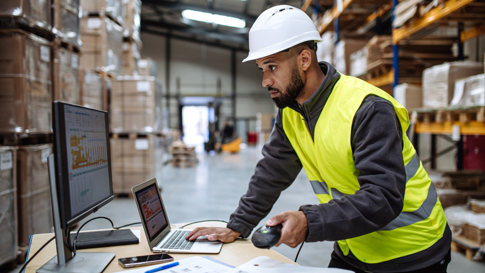 An image of a warehouse worker looking at data on a computer in a warehouse environment. He is wearing a hard-hat and a high-visibility vest, while holding a scanner in his left hand.