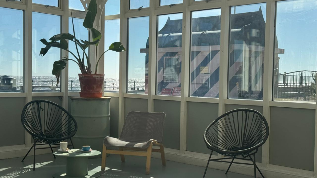 An image of the interior of the East Point Pavilion, featuring chairs and a potted plant near a glass window. Outside of the window, the seafront can be seen.