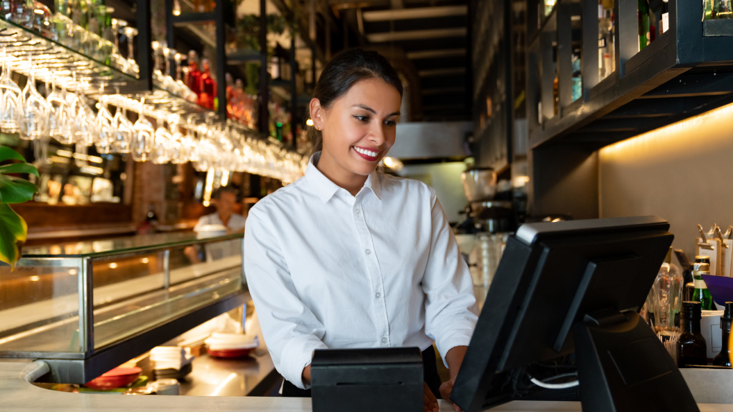 An image of a Front of House Manager using a computer system in a restaurant. She is in the bar area and wearing a smart shirt.