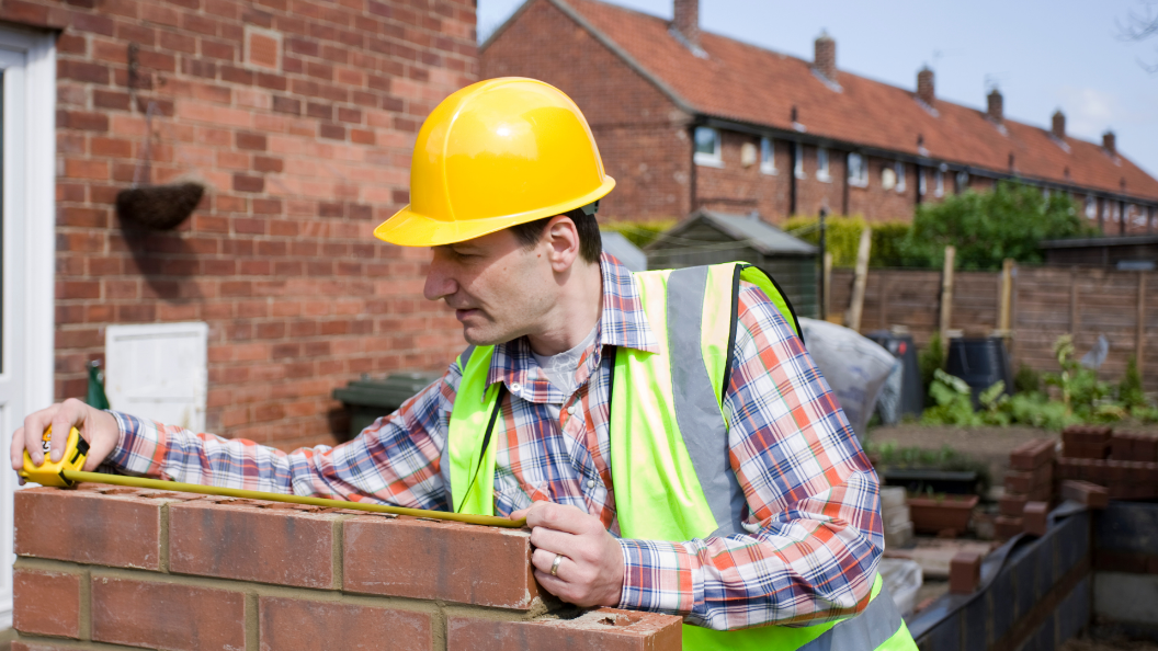 An image of a bricklayer measuring a wall in a garden. He is wearing a yellow hard-hat and a high-visibility vest