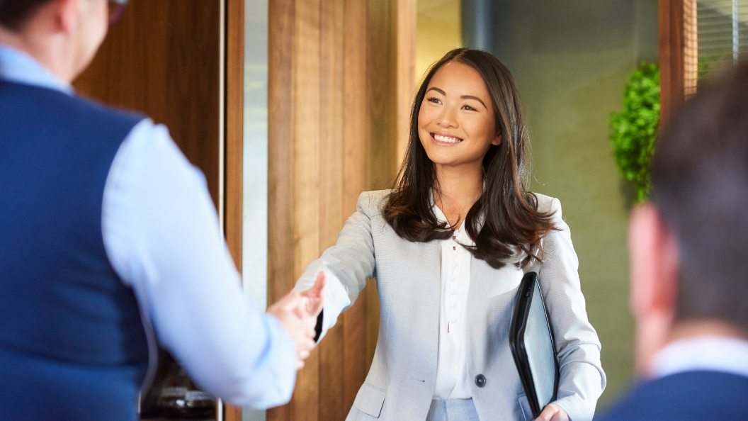 An image of a woman shaking hands with a potential employer. She is standing and smiling, while the two people across from her are sat and viewed from the back