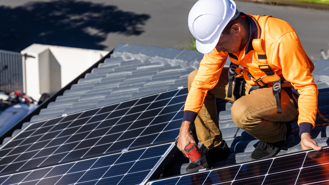 An image of a worker installing a solar panel on a roof, wearing safety equipment.