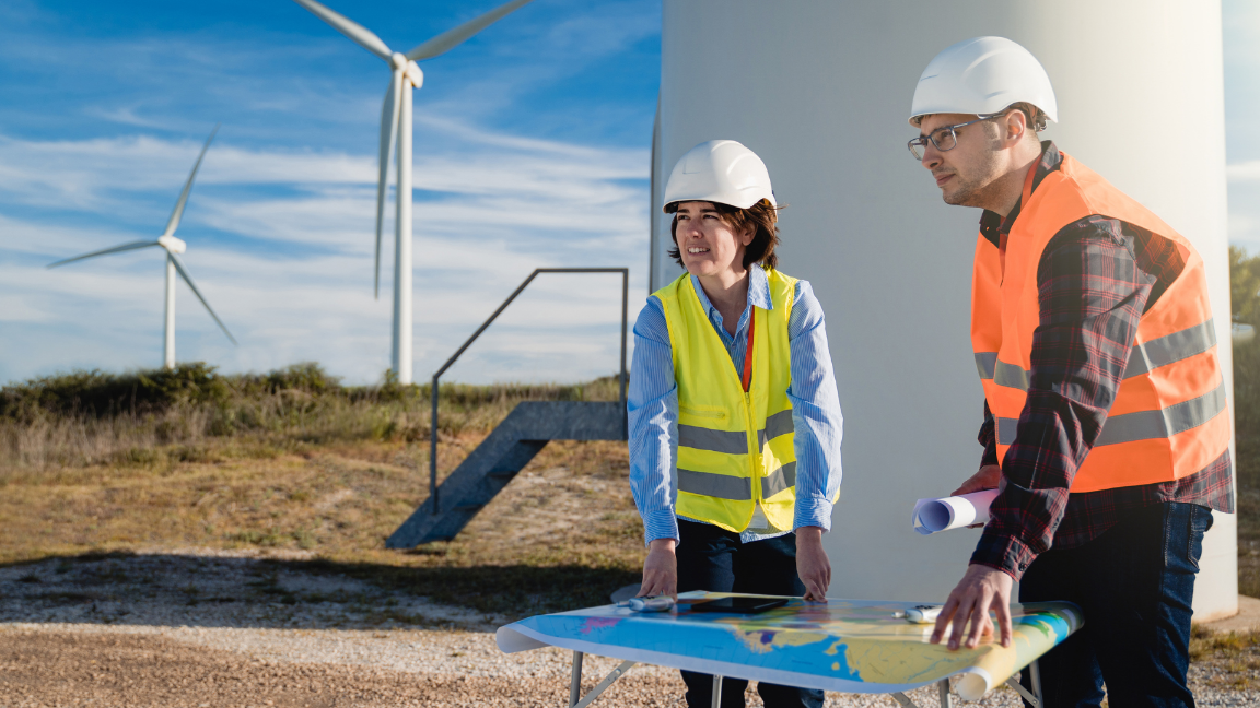 Two people looking at a map on a table on a hillside. They are wearing high-visibility vests and hard hats. There are wind turbines on the hill behind them.