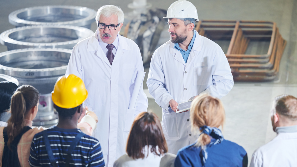 An image of a staff meeting in a factory. There is a manager and a supervisor addressing a group of employees.