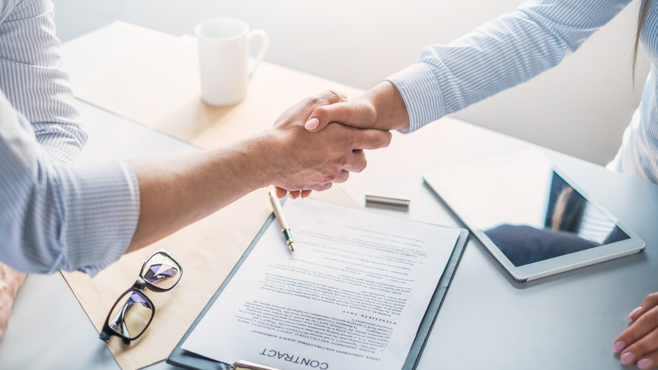 An image of two people shaking hands over a contract on a desk.