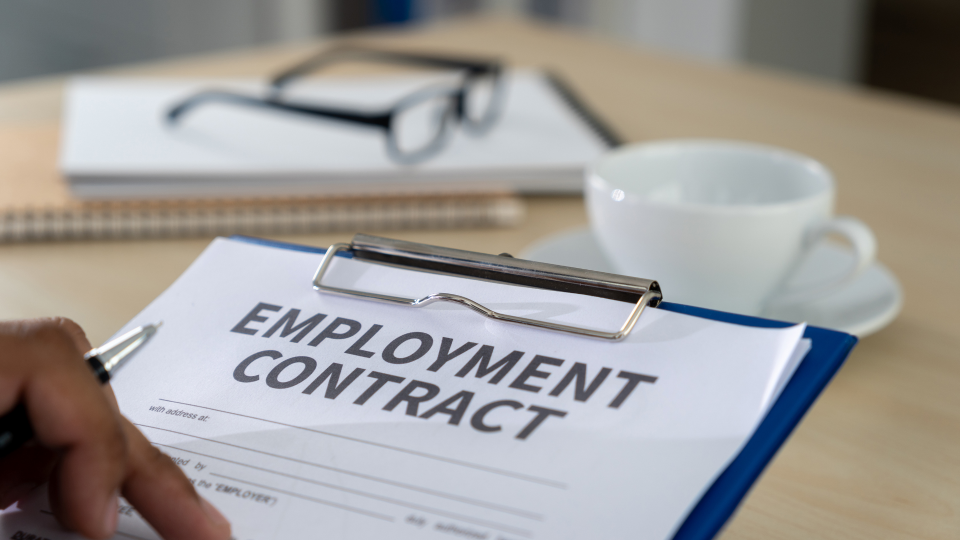 An image of an employment contract on a blue clipboard. The clipboard is on a wooden desk, with a coffee cup and a notepad.