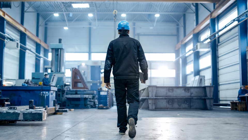 An image of a Production Manager walking through a factory with a clipboard in his hand. There are large metal components all around the factory.