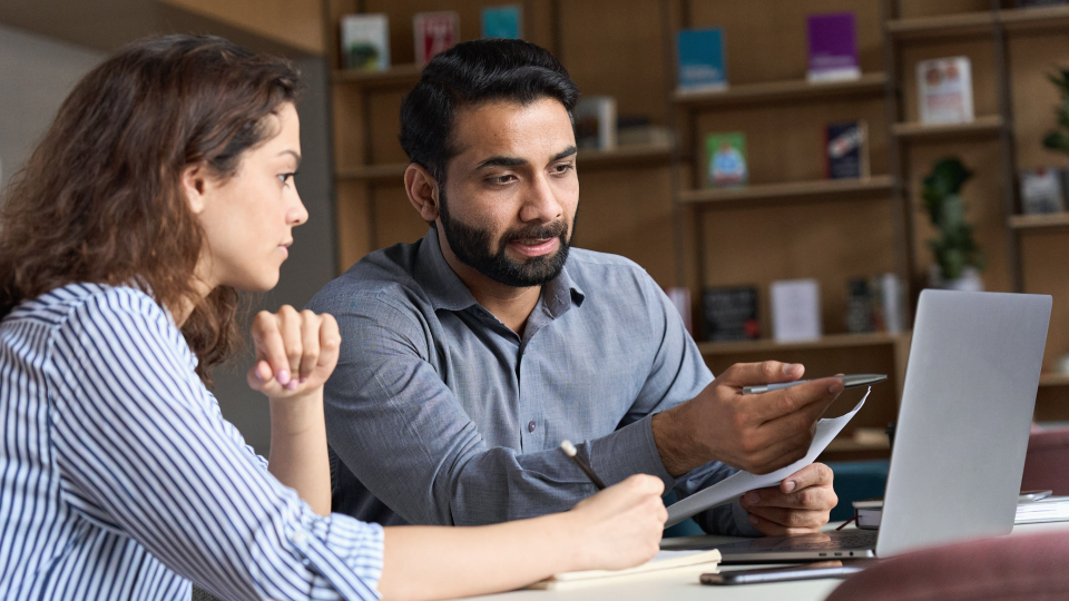 An image of two people discussing something on a laptop. The man on the left is holding a pen and a piece of paper, and pointing at something on the screen. The woman on the right is writing in a notepad.