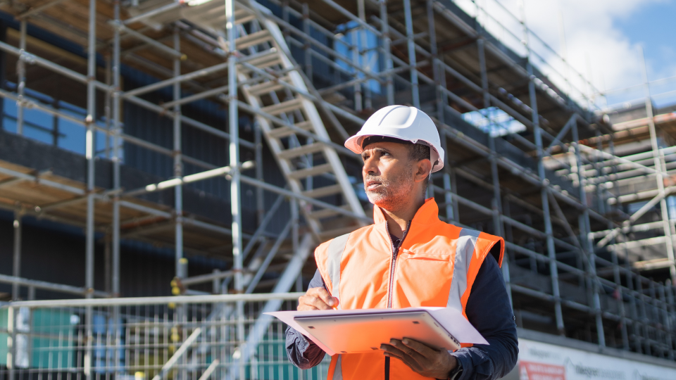 An image of an architect visiting a construction site. He is wearing a white hardhat and an orange high-visibility jacket, while holding a laptop and a piece of paper.