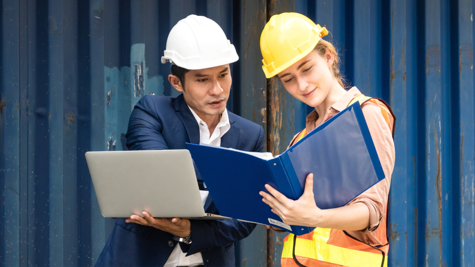 An image of two workers discussing an engineering project. One person is wearing a suit and is holding a laptop. The other is wearing high-visibility wear and is holding a folder.