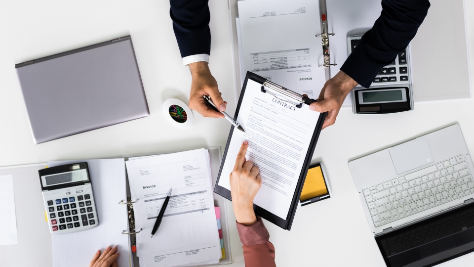 An top-down image of two people discussing financial reports on a white desk. They are sat opposite each other and both have copies of the reports and a calculator.
