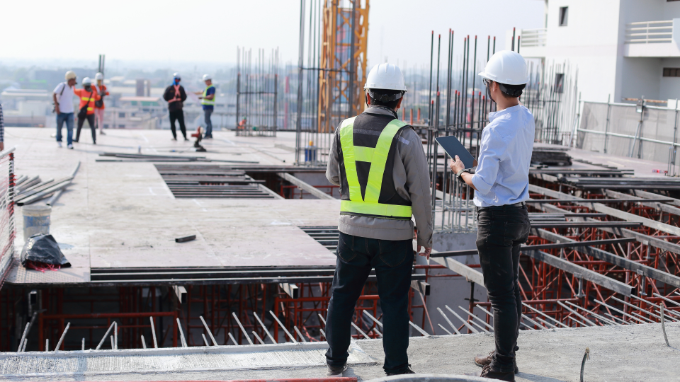 An image of two workers on top of a building under construction. They are looking at data on a tablet and reviewing the structure of the building.