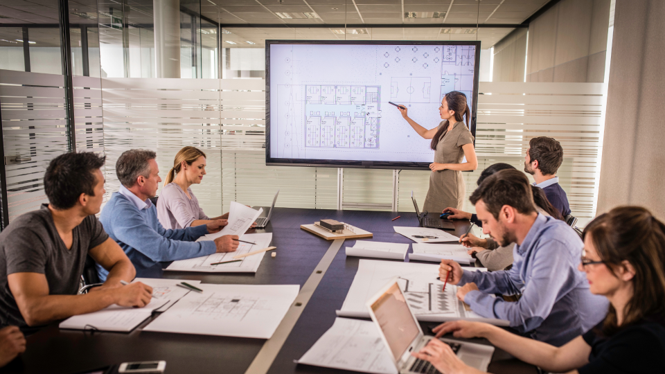 An image of eight people discussing an architectural drawing on a large display in a meeting room. One woman is stood up next to the display and pointing at parts of the drawing.