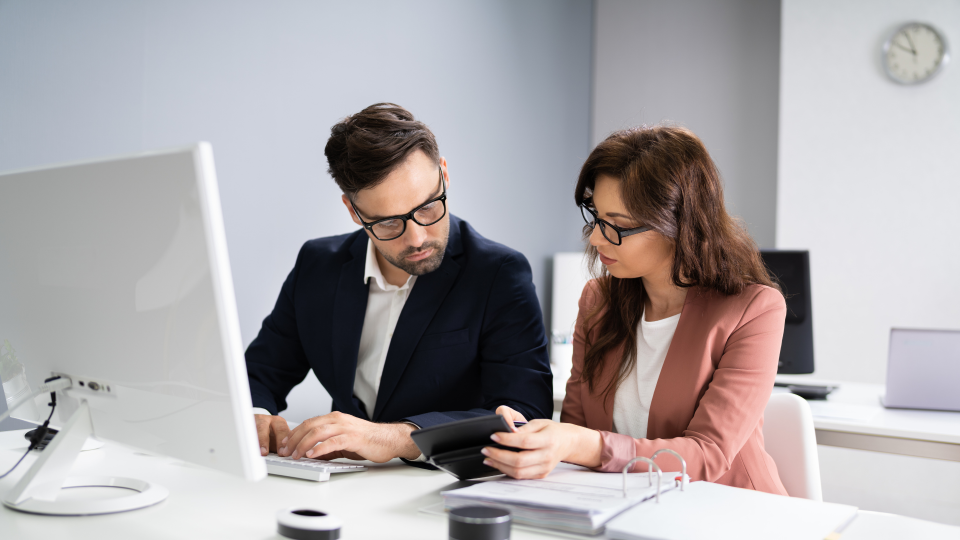 An image of two people looking at financial data. They are sat next to each other at a white desk. There is a white computer monitor on the desk and a folder full of reports. Both people are looking at a calculator.