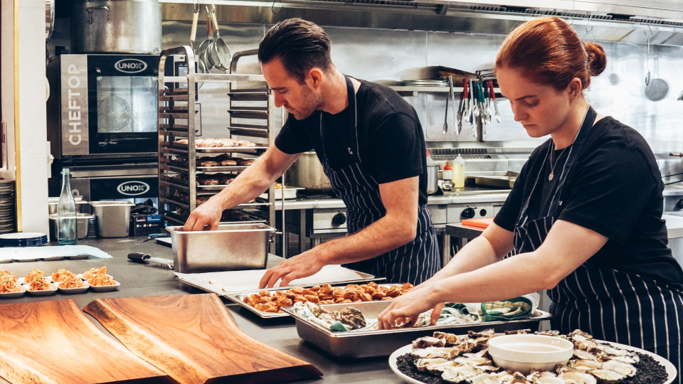 An image of two chef plating ingredients in a commercial kitchen. They are wearing black t-shirts and blue aprons, and there is commercial cooking equipment behind them.