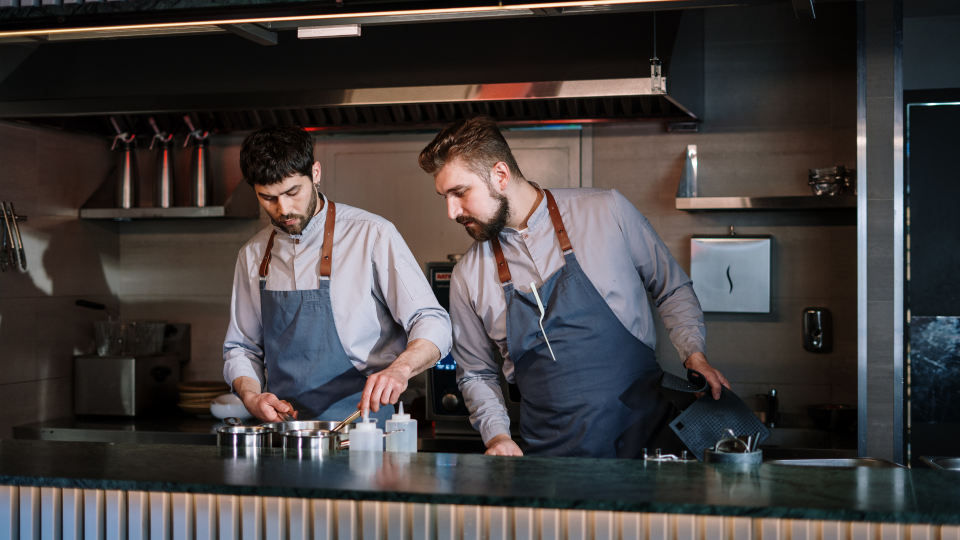 An image of two chefs in a kitchen. One chef is cooking in a saucepan while the other chef is checking what is being made.