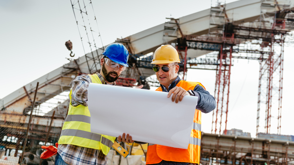 An image of two construction workers looking at technical drawings for an infrastructure project. There is a bridge under construction behind them.