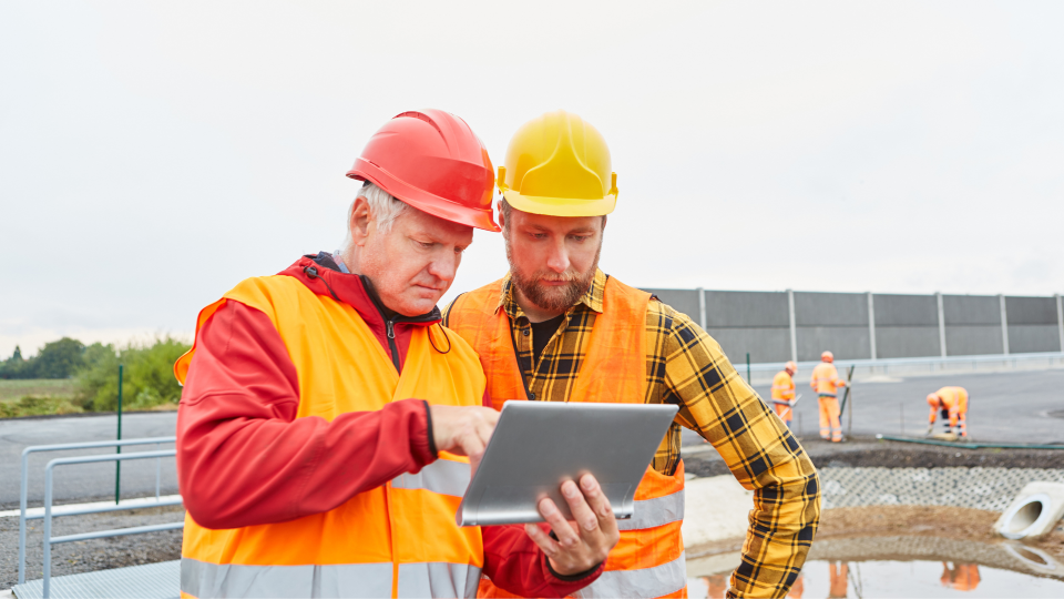 An image of two construction workers on a site looking at a digital tablet. They are wearing hard hats and high-visibility vests