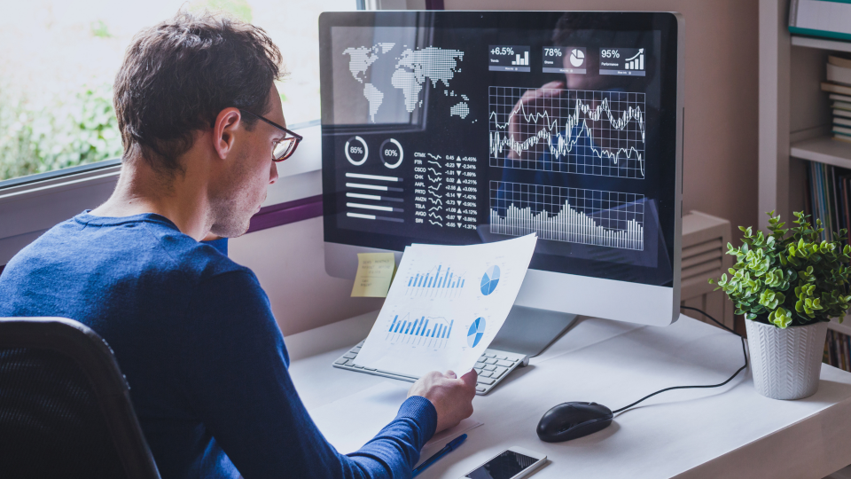 An image of a man looking at financial data on both a piece of paper and a computer screen. He is sat at a small white desk next to a window.