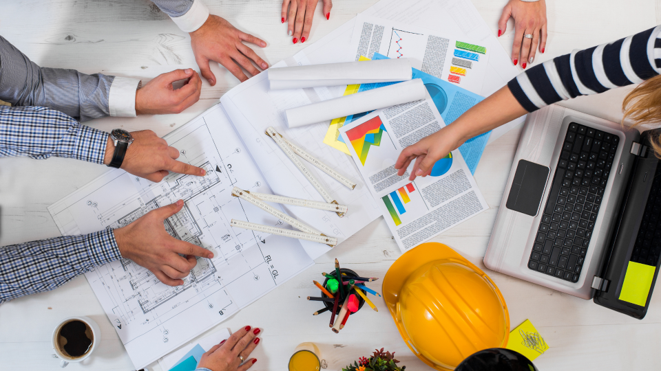 A top-down image of five construction workers discussing a project. There are building blueprints and costing sheets on the desk.