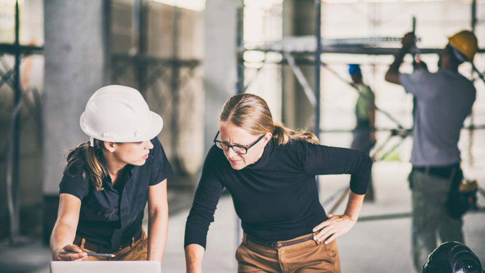 An image of two women looking at a laptop on a construction site. There are construction workers carrying materials in the background