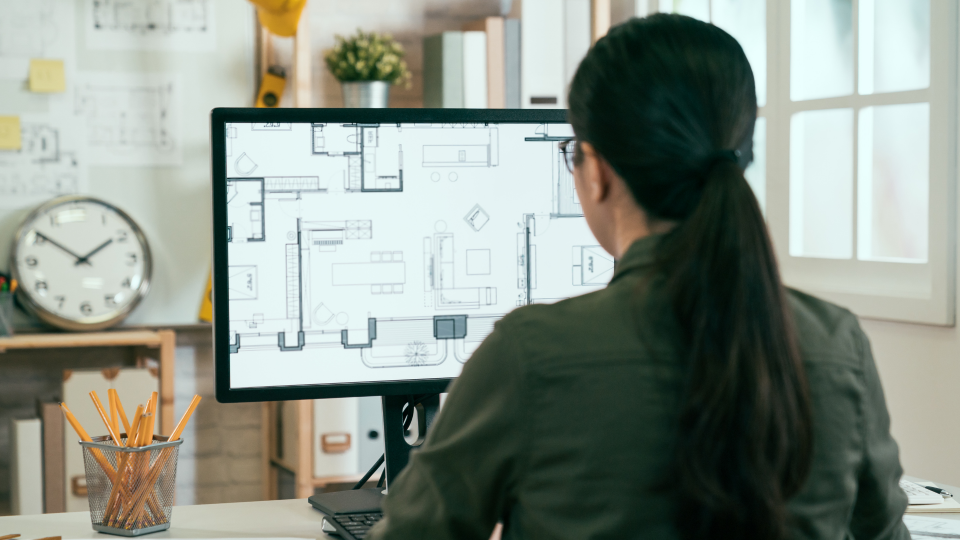 An image of a woman looking at a digital plan of a building. She is sat at a desk and is looking at a single computer screen