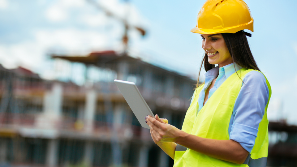 An image of a woman looking at a tablet device on a construction sire. She is wearing a yellow hard-hat and a high-visibility vest.