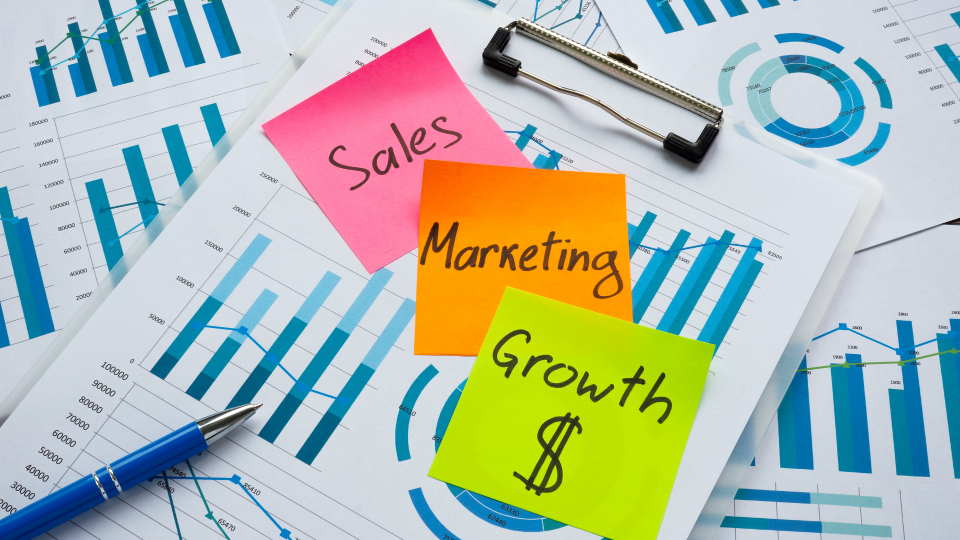An image of several charts on pieces of paper. The top piece of paper has three post-it notes on it, saying Sales, Marketing, and Growth