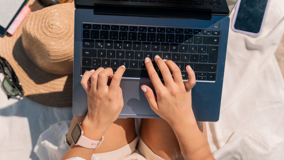 An image of a woman using a laptop. She is sat outside on a sheet, with her phone and sunglasses next to her