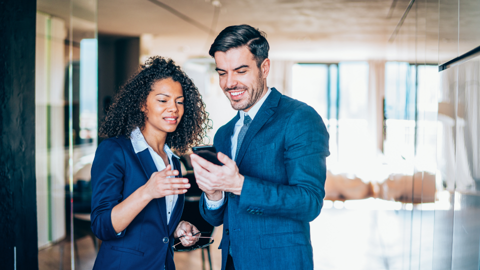 An image of a man and a woman looking at a mobile phone. They are standing up in an office and are both wearing a blue suit