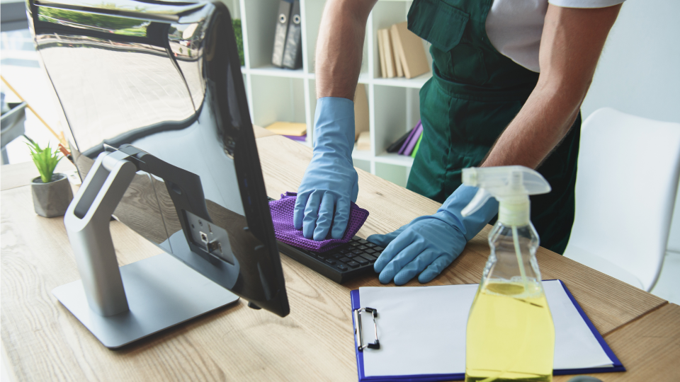 An image of a cleaner cleaning a computer keyboard on an office desk. The cleaner is wearing blue gloves and a green apron, and is using a purple cleaning-cloth.