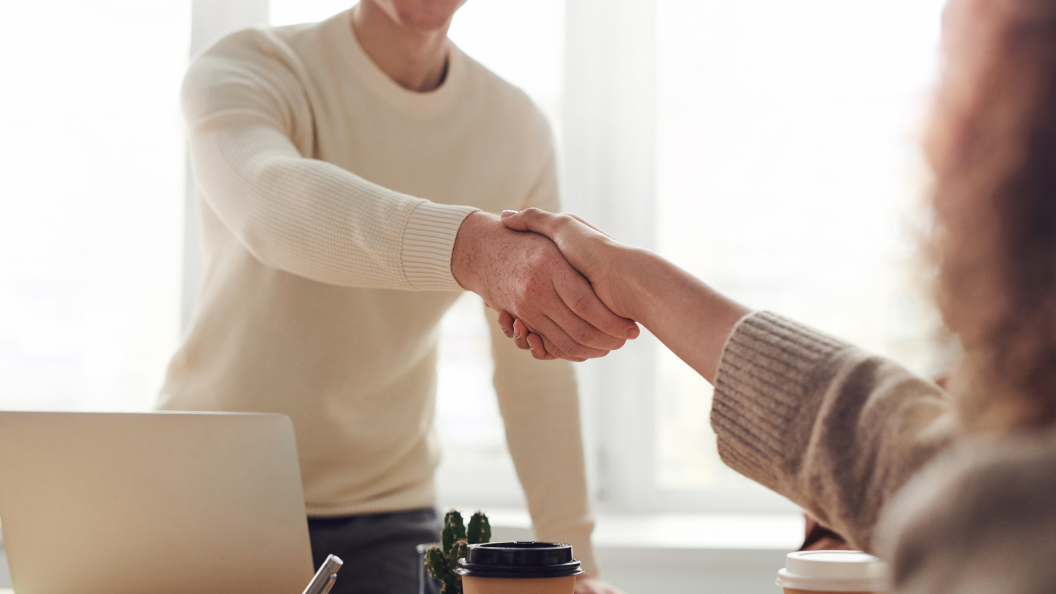 An image of two people shaking hands over a desk. The person furthest from the camera is standing up, and has a laptop facing towards them.
