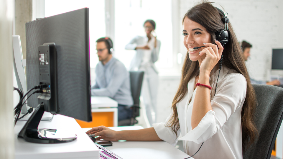 An image of several customer service workers in an office. They are sat at individual desks and are wearing headsets plugged in to computers